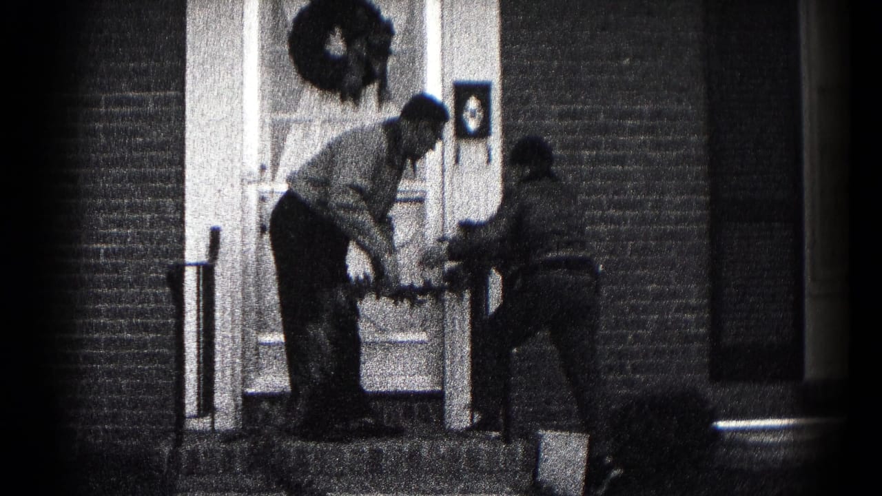 Two people stand at a brick home's front steps while handling a Christmas wreath near the door.