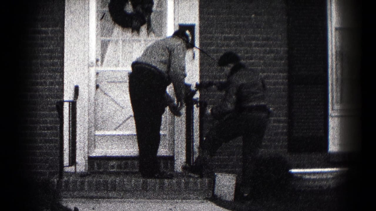 Two men work near the front steps of a brick house with a wreath on the door.