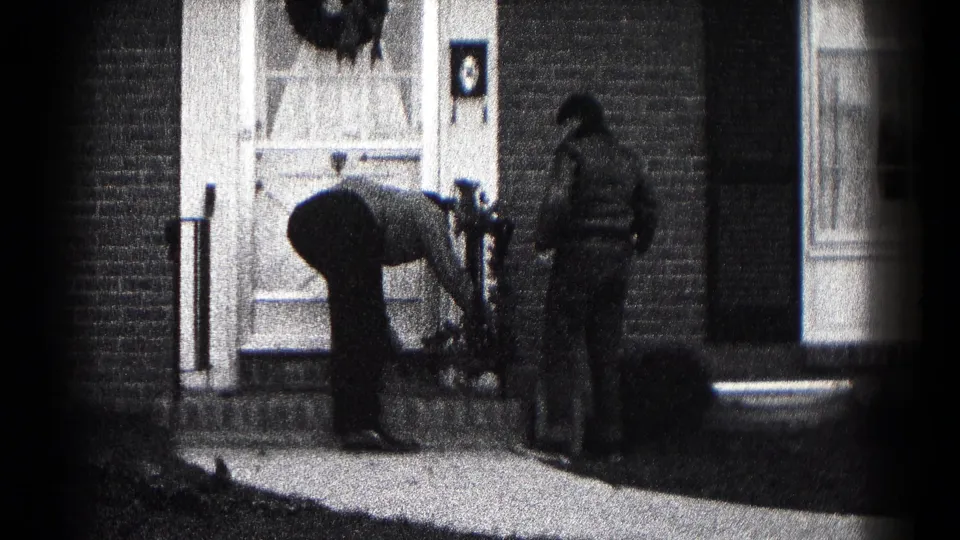 Black-and-white 8mm frame of two men arranging Christmas decor outside a Hagerstown brick residence in 1959.