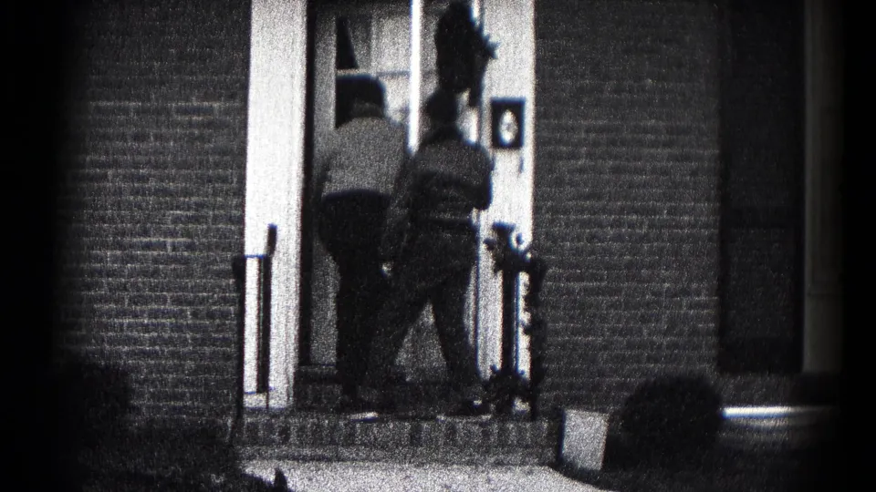 The men move closer to the entrance of the brick home, with the door area and stoop railings visible in the frame.