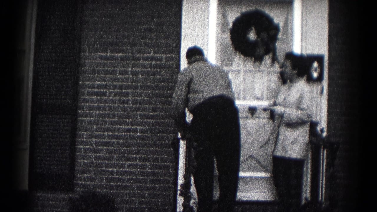 1959 Hagerstown home-movie frame showing two adults at a wreath-decorated front door during a farewell.