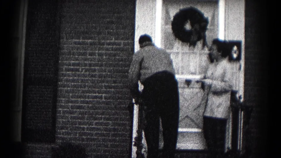 1959 Hagerstown home-movie frame showing two adults at a wreath-decorated front door during a farewell.