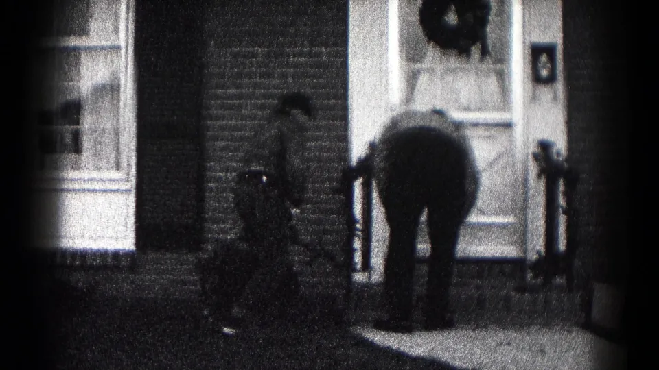Black-and-white 8mm frame from Hagerstown shows one person bent at the porch and another nearby at a brick house entrance.