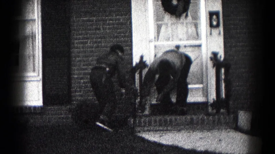 The final frame shows a figure bent low at the porch with another nearby, the wreath still visible on the front door.