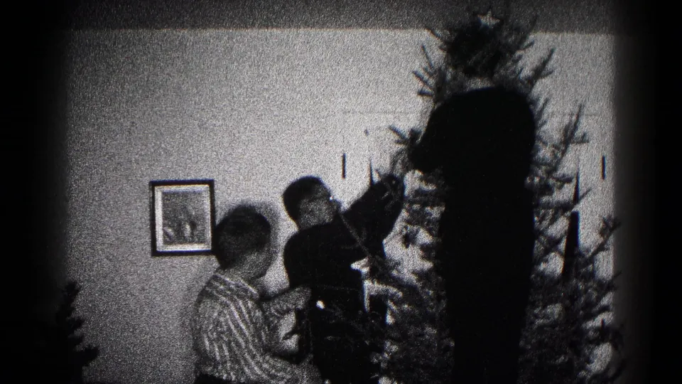 Grainy 8mm frame from 1959 Hagerstown showing adults and a child gathered at a Christmas tree in a private room.