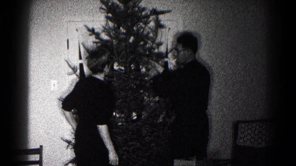 Two people appear to adjust ornaments or lights on a Christmas tree in a grainy 1959 Hagerstown home-movie interior.