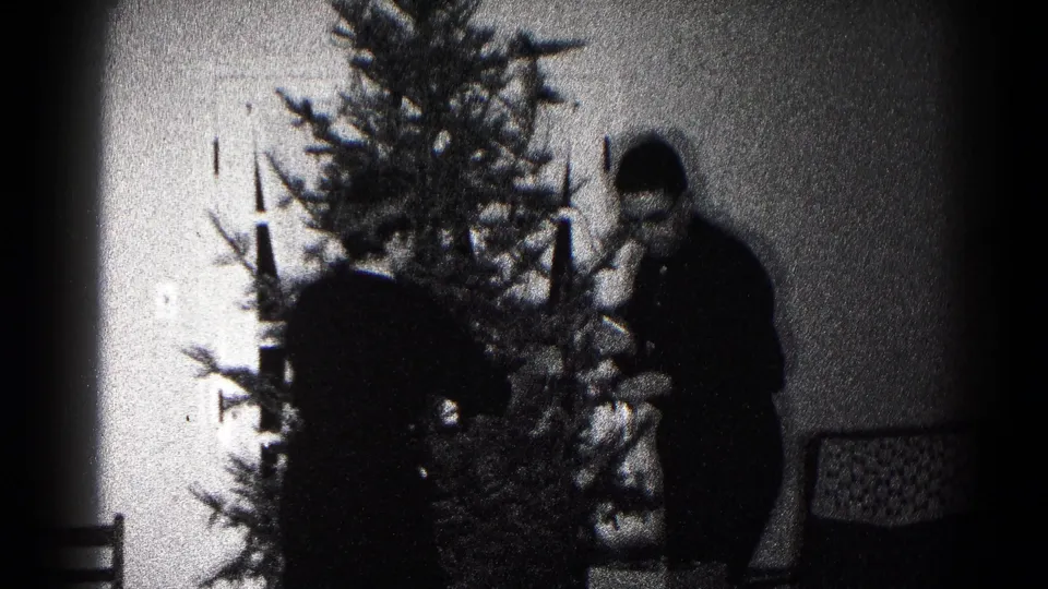 In a 1959 Hagerstown home-movie frame, adults stand near a Christmas tree while decorating inside a living room.