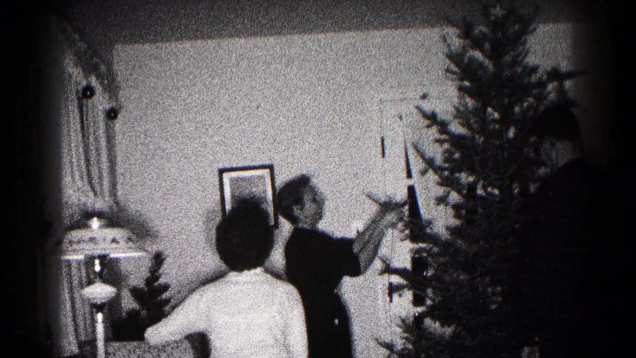 Black-and-white 1959 Hagerstown home-movie frame showing people beside a Christmas tree, with a lamp and framed picture in the room.