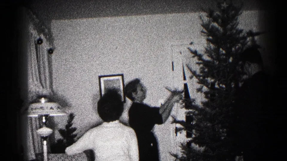 Black-and-white 1959 Hagerstown home-movie frame showing people beside a Christmas tree, with a lamp and framed picture in the room.