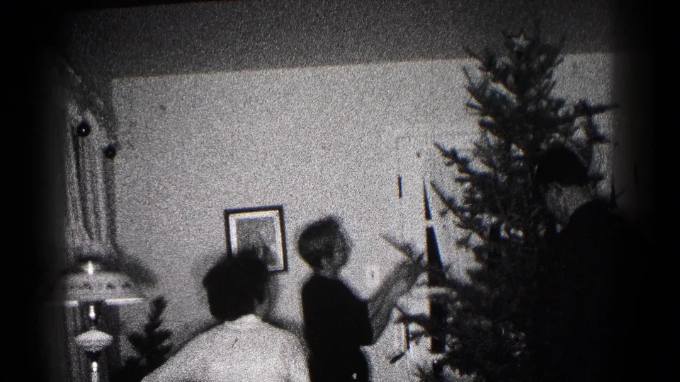 Black-and-white 1959 Hagerstown home-movie frame with a Christmas tree, seated figure, lamp, wall picture, and decorator at right.