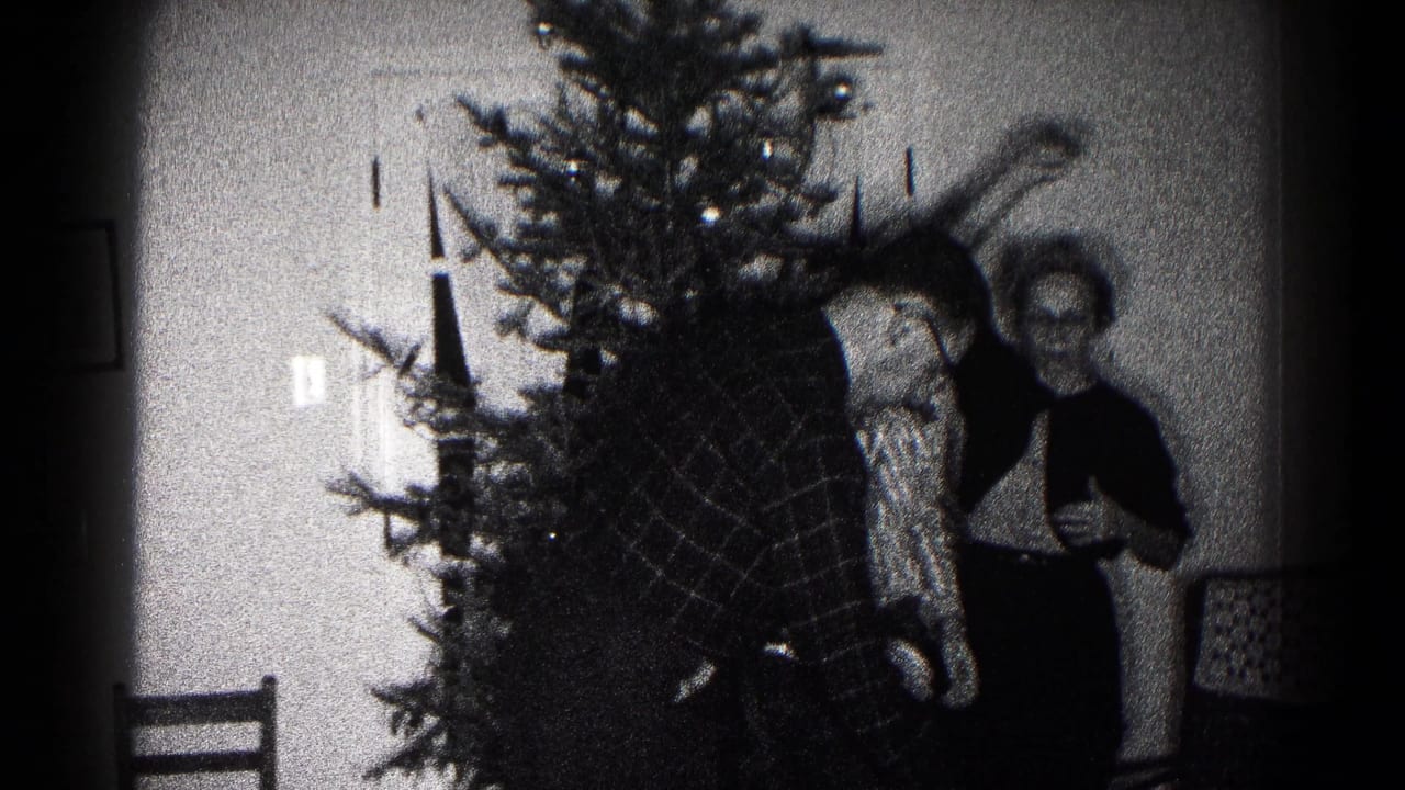 Three people stand near a Christmas tree inside a room with chairs and a plain door.