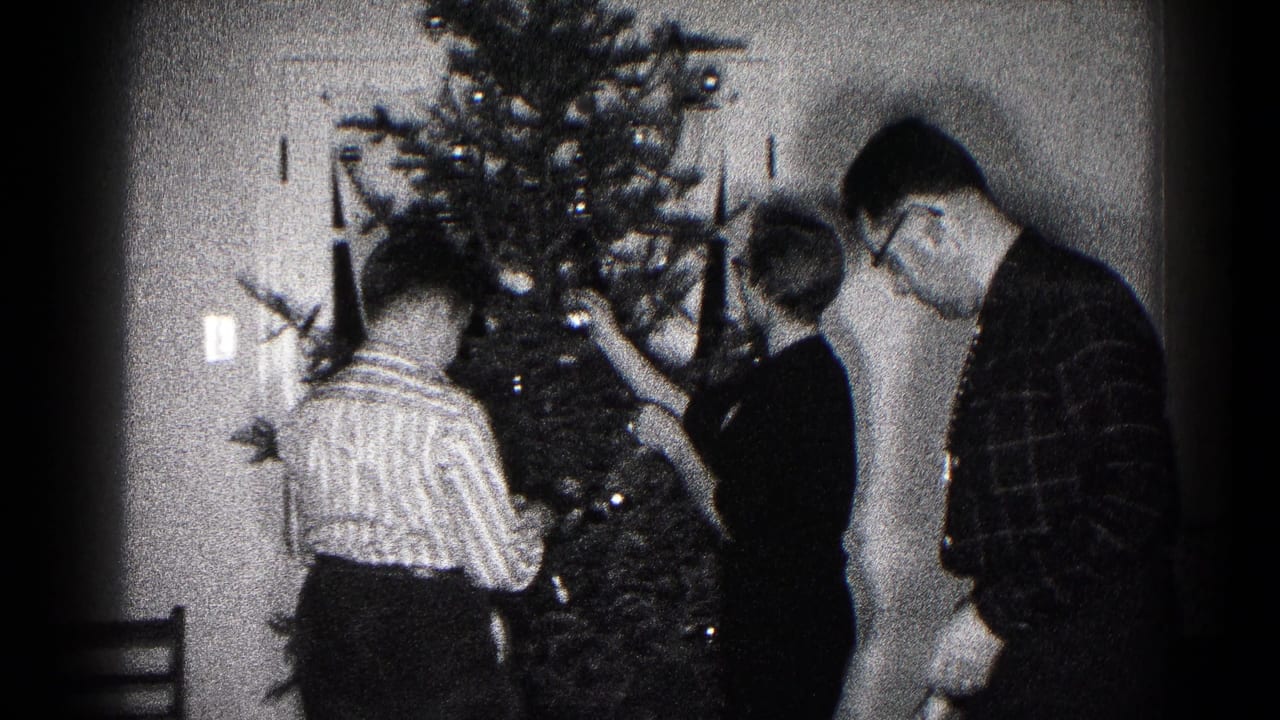 Three people hang ornaments on a Christmas tree near a plain door.