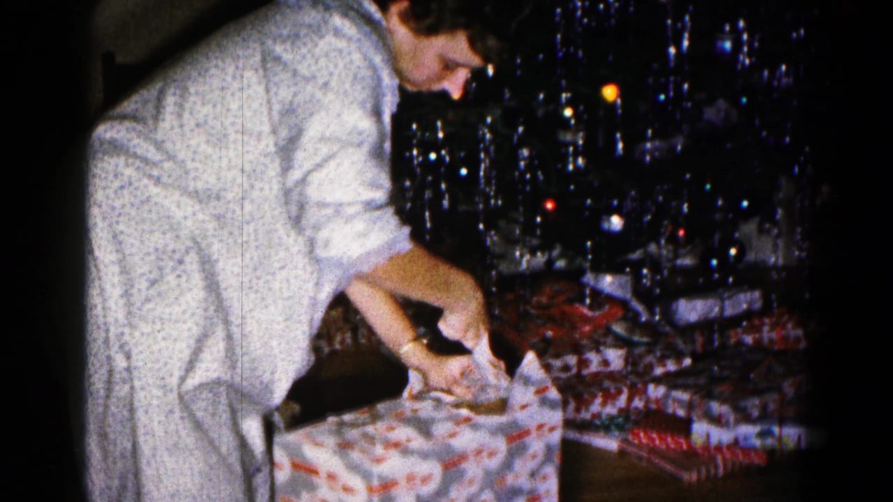 Woman in light pajamas unwraps a patterned Christmas present beside a decorated tree.