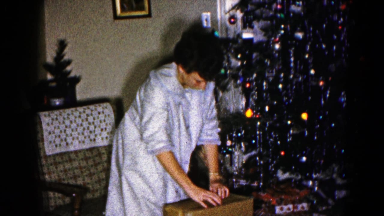 In the 1959 Hagerstown living room, a woman in light pajamas leans over a cardboard box beside the Christmas tree and gifts.