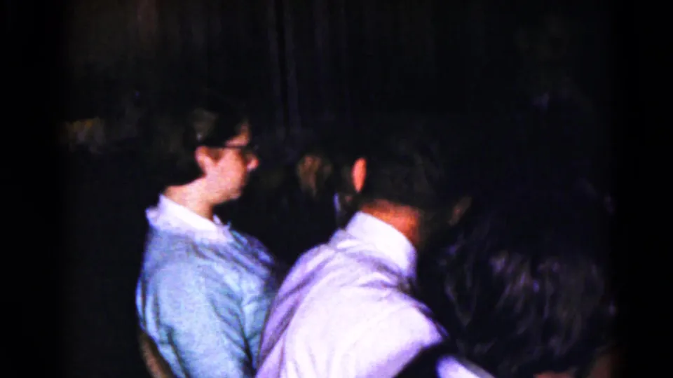 1959 8mm frame showing two seated adults in dim light, one in profile with glasses and another turned away in a light shirt.