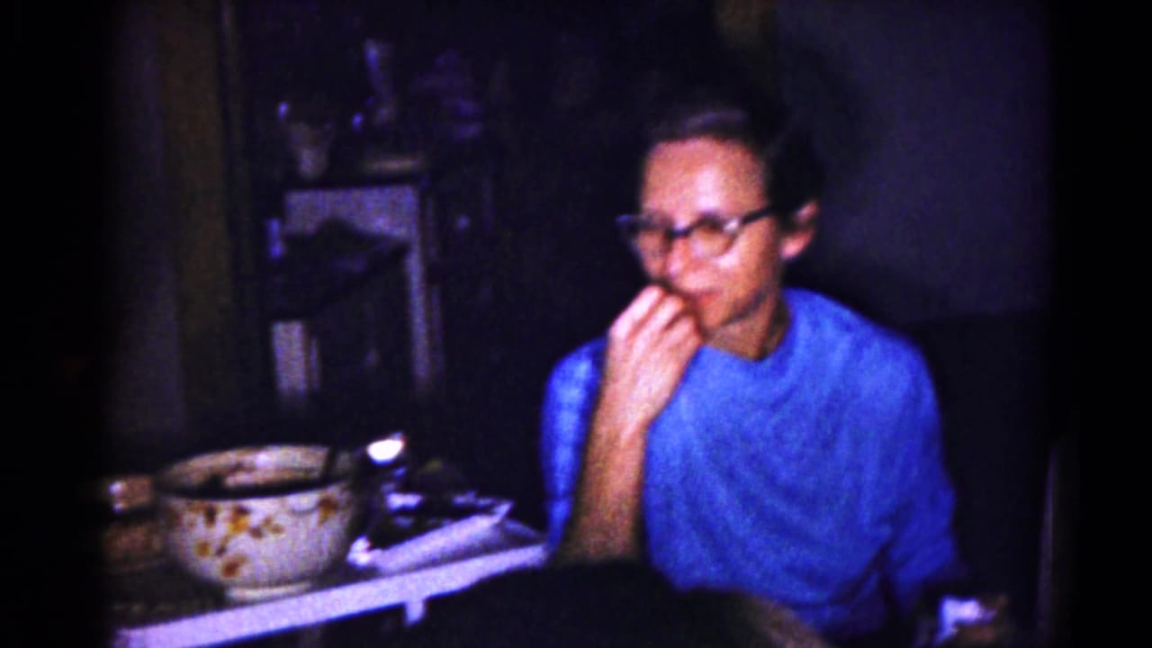 In a 1959 Hagerstown home-movie frame, a man in glasses and a blue shirt eats beside tableware in a dark interior.