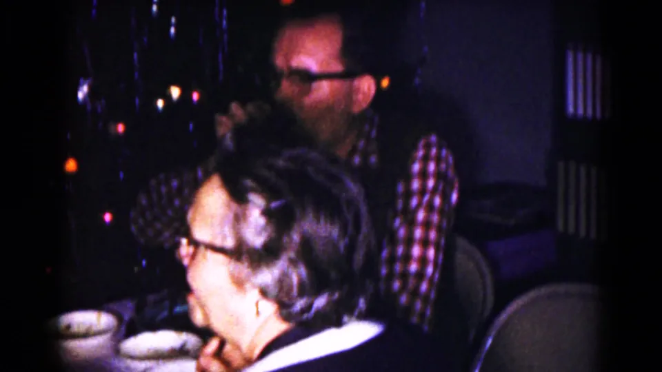 A woman in glasses sits in the foreground while a man in plaid stands or sits behind her near holiday lights in the private home.