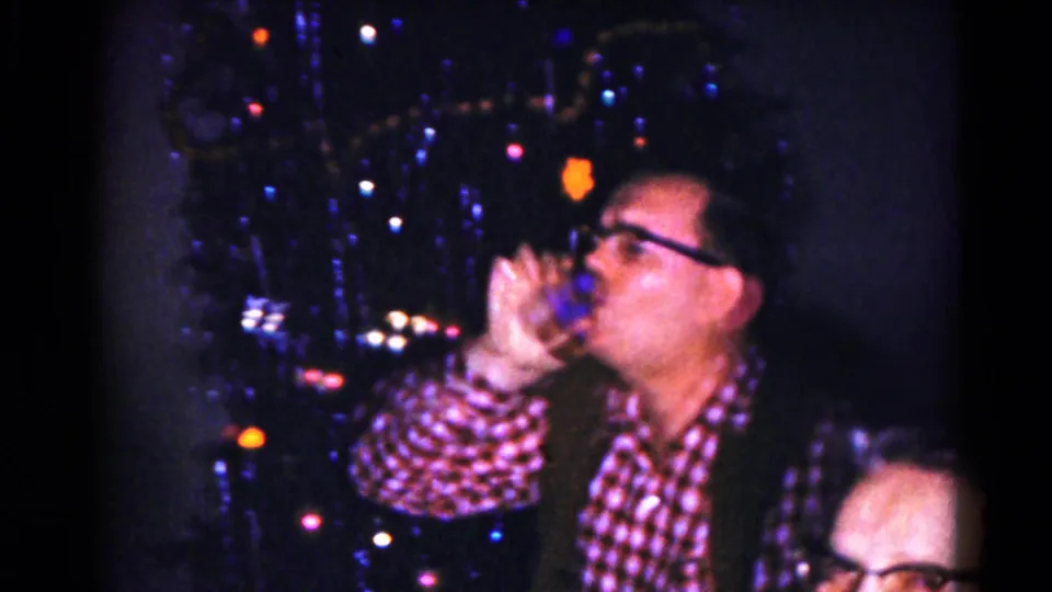 A man in a plaid shirt and glasses drinks from a glass next to the lit Christmas tree during the 1959 family dinner scene.