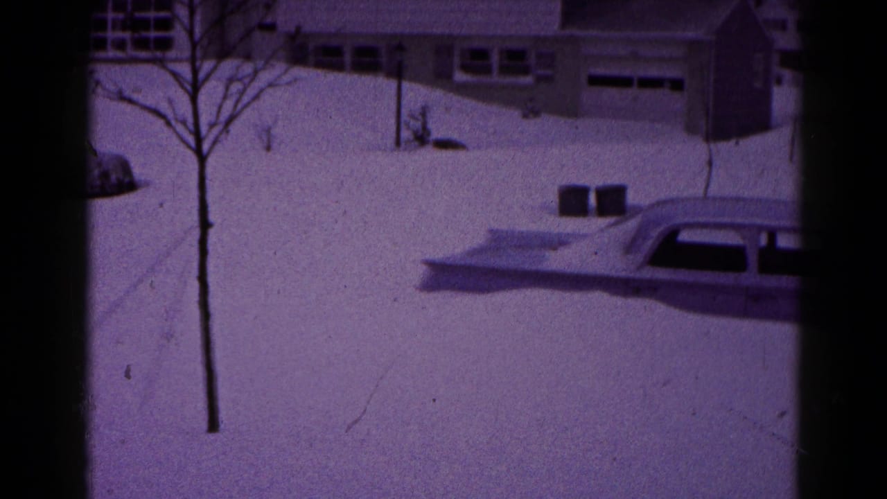 1959 Hagerstown home-movie frame showing a snowbound suburban street, a partially buried sedan, bare tree, and house with garage in the background.
