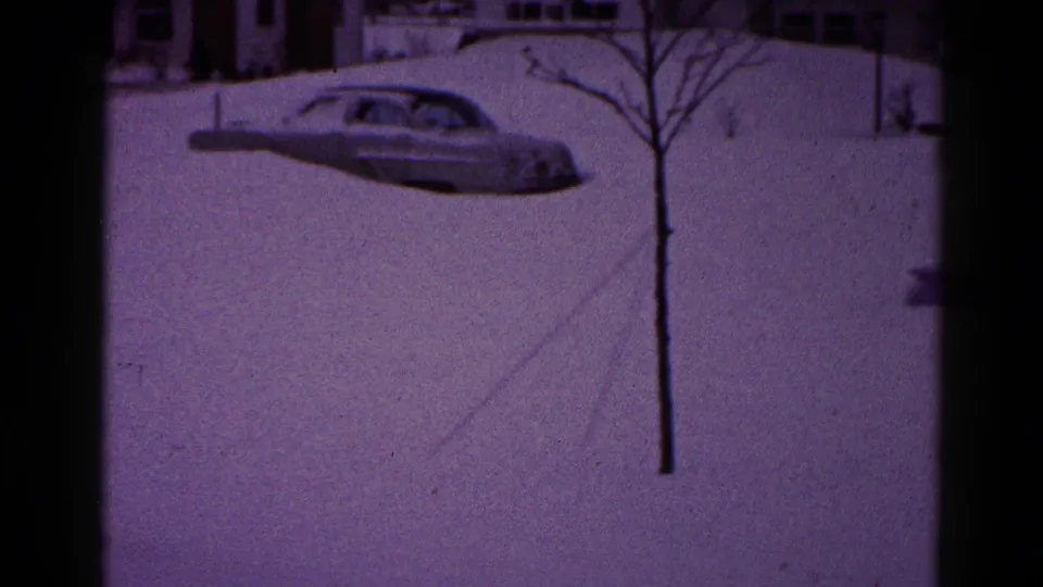 A 1959 Hagerstown street frame shows a rounded American sedan half-buried in snow, with bare trees and residential buildings behind it.
