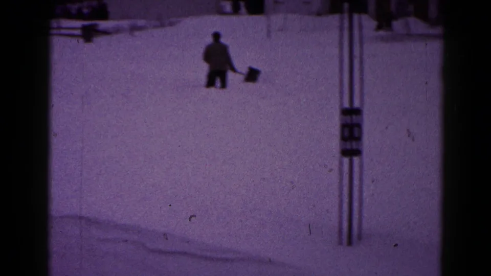 1959 Hagerstown frame showing a bundled man shoveling or carrying snow near utility poles in a snow-covered residential area.