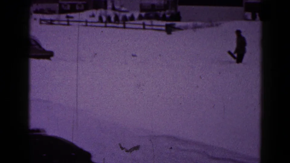 1959 Hagerstown home-movie frame with a distant man carrying a shovel across deep snow near a fence and residential roadside.