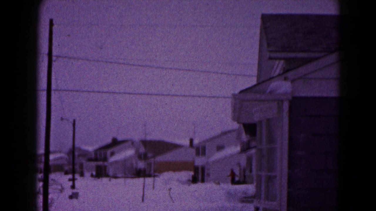 Snowy neighborhood street with houses, utility poles, overhead wires, and deep snowbanks.