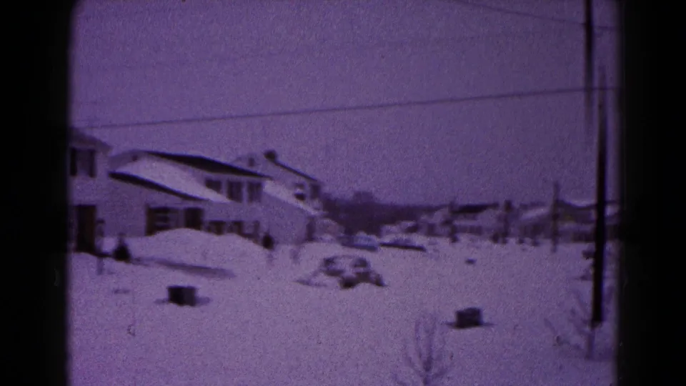 1959 Hagerstown winter neighborhood frame showing houses, overhead wires, heavy snow, and a partly buried vehicle or snow-covered mound.