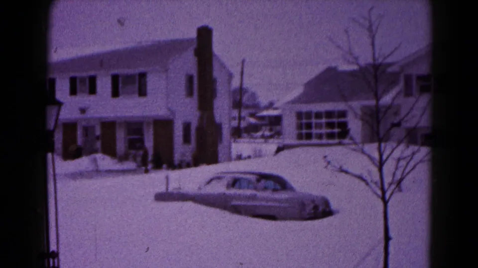 1959 Hagerstown frame of a snow-covered suburban block with house fronts, chimney, bare tree, and a partly buried rounded car.