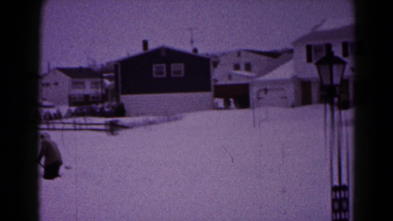 1959 Hagerstown home-movie frame showing deep snow across residential yards, houses, fencing, and a distant person near the left edge.