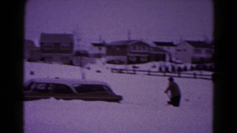 1959 Hagerstown winter frame with a person shoveling near a car buried in deep snow outside suburban homes.