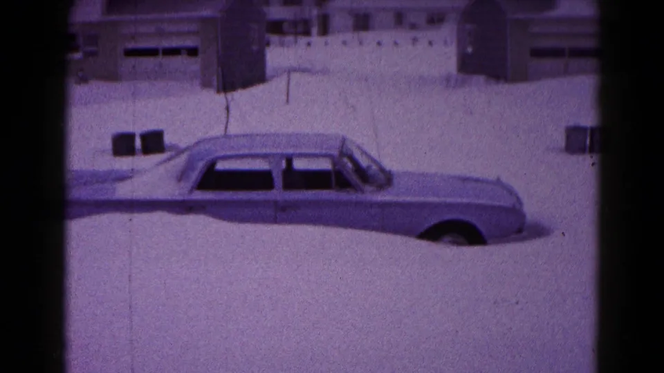 1959 Hagerstown home-movie view of a large American car partly buried by snow near houses and a driveway or street edge.