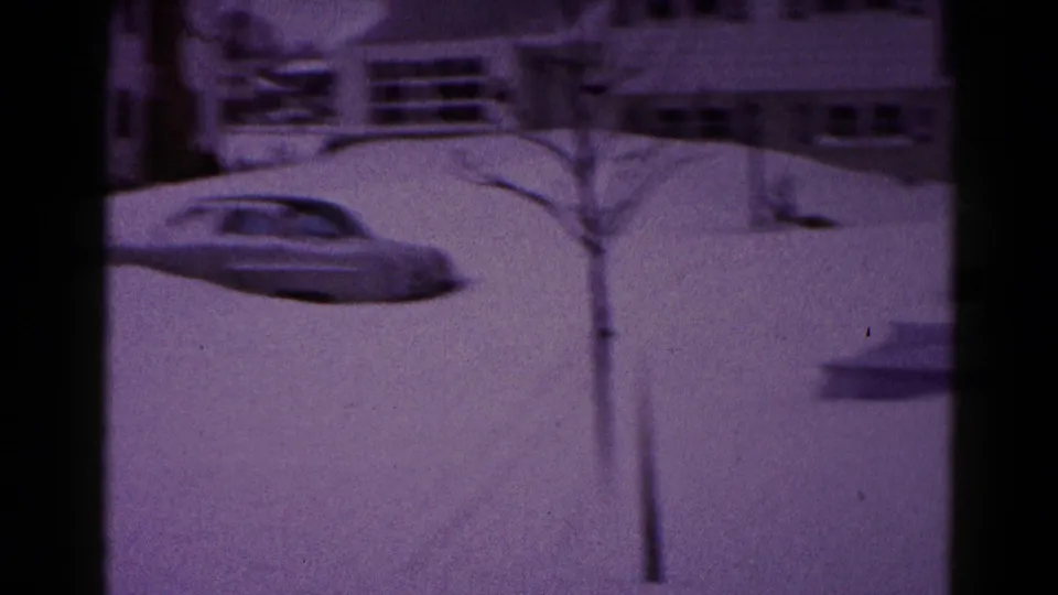 1959 Hagerstown winter frame showing a snowbound car, a small bare tree, deep snowbanks, and residential houses in the background.