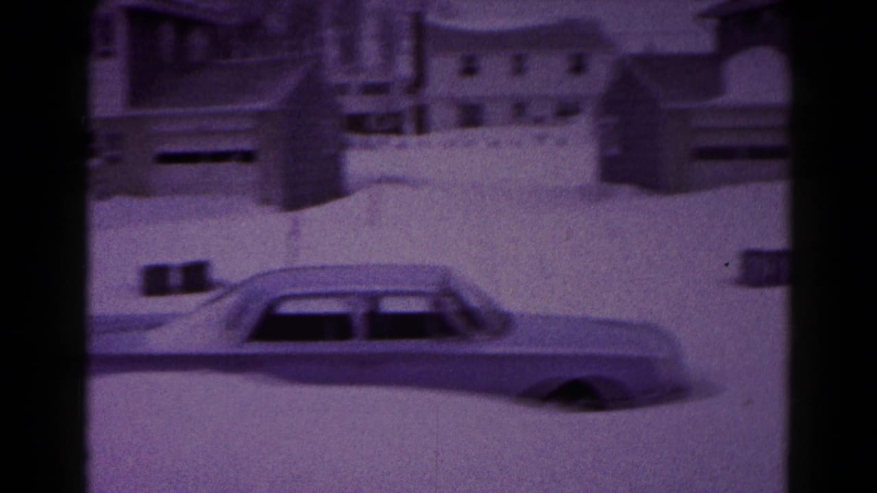 Large car partly buried in snow in front of houses and snowbanks.
