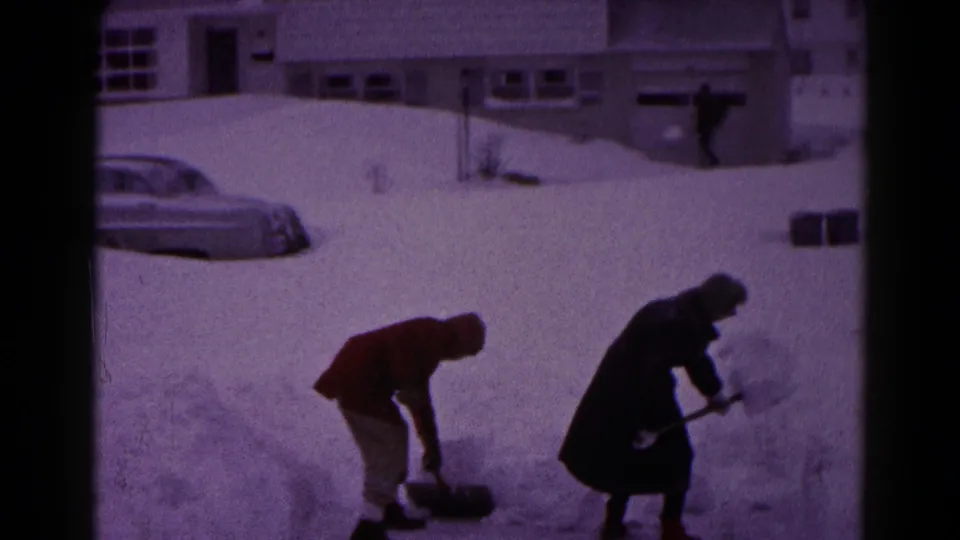 In 1959 Hagerstown, Maryland, two bundled people shovel snow in a residential yard with a snow-covered car and houses behind them.