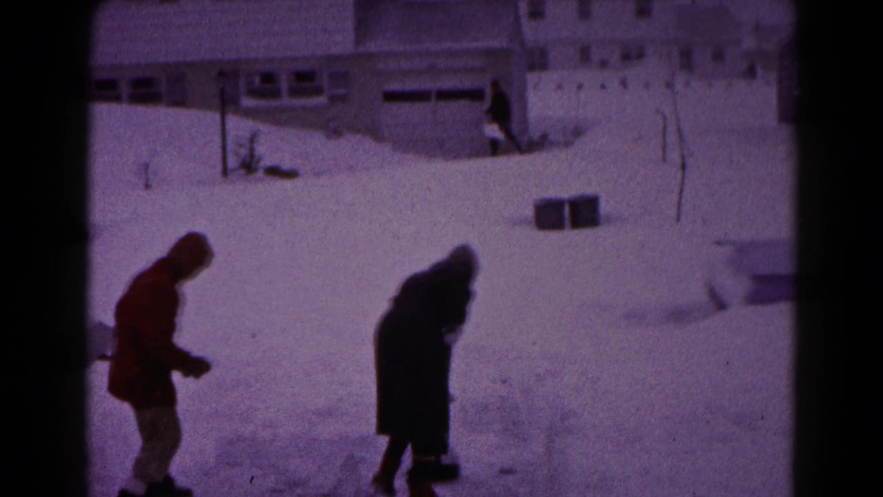 Two bundled figures stand in deep snow near a residential house and snowbanks.