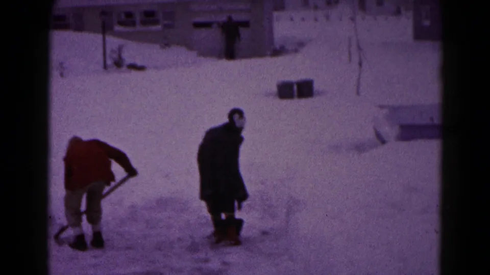 Two bundled adults shovel and walk through deep snow in a Hagerstown residential neighborhood, with houses behind them.