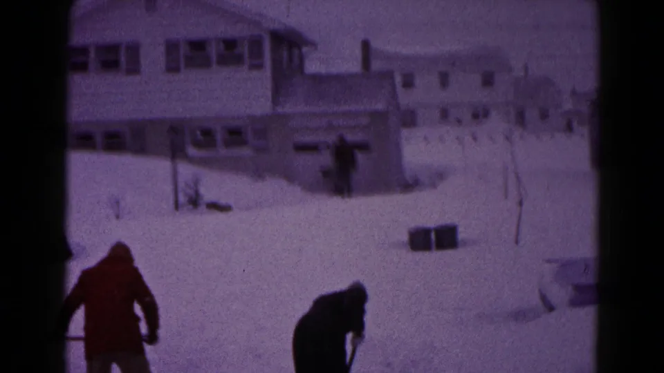The home-movie frame shows two men working low over their shovels while snow covers the ground and nearby houses.
