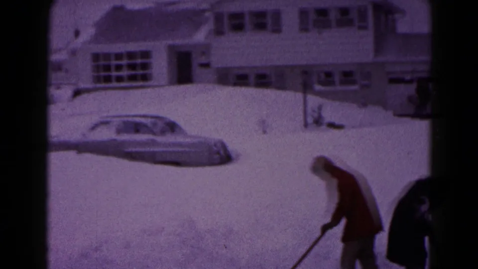 A man in a red coat shovels snow near a partly buried mid-century car outside a residential house in Hagerstown.