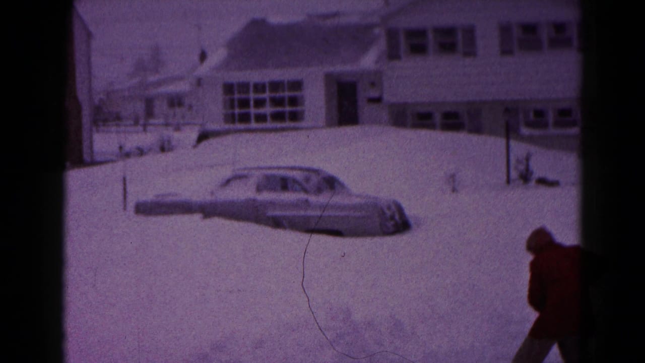 1959 Hagerstown home-movie frame showing a snow-covered suburban house, a grey sedan in deep snow and a person in a red coat at the edge of the scene.