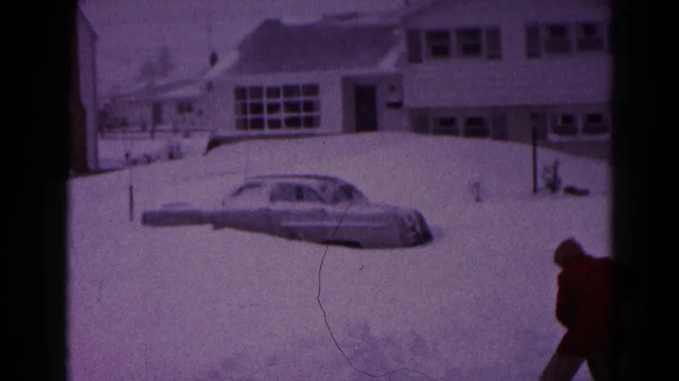 1959 Hagerstown home-movie frame showing a snow-covered suburban house, a grey sedan in deep snow and a person in a red coat at the edge of the scene.