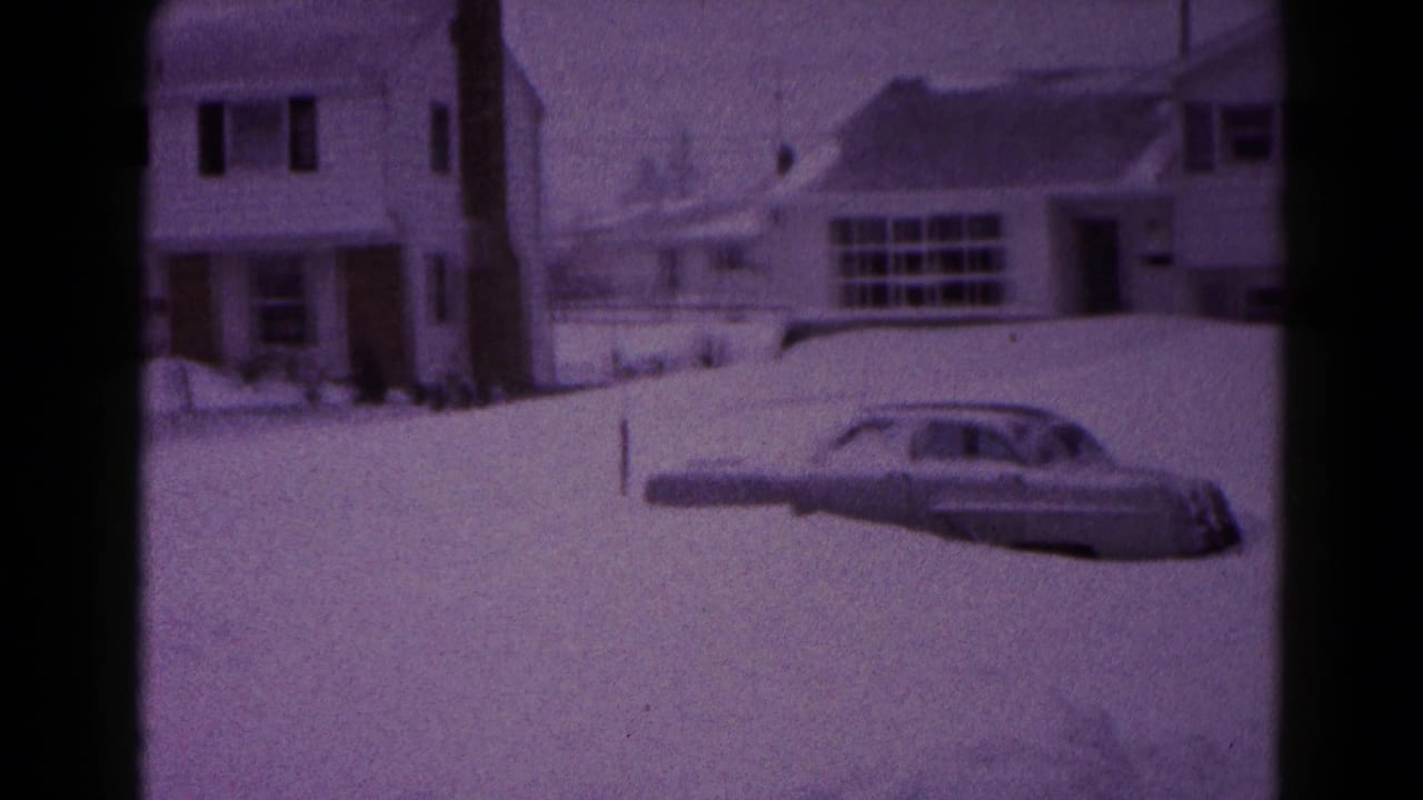 Grey car partly buried in snow beside snow-covered suburban houses.
