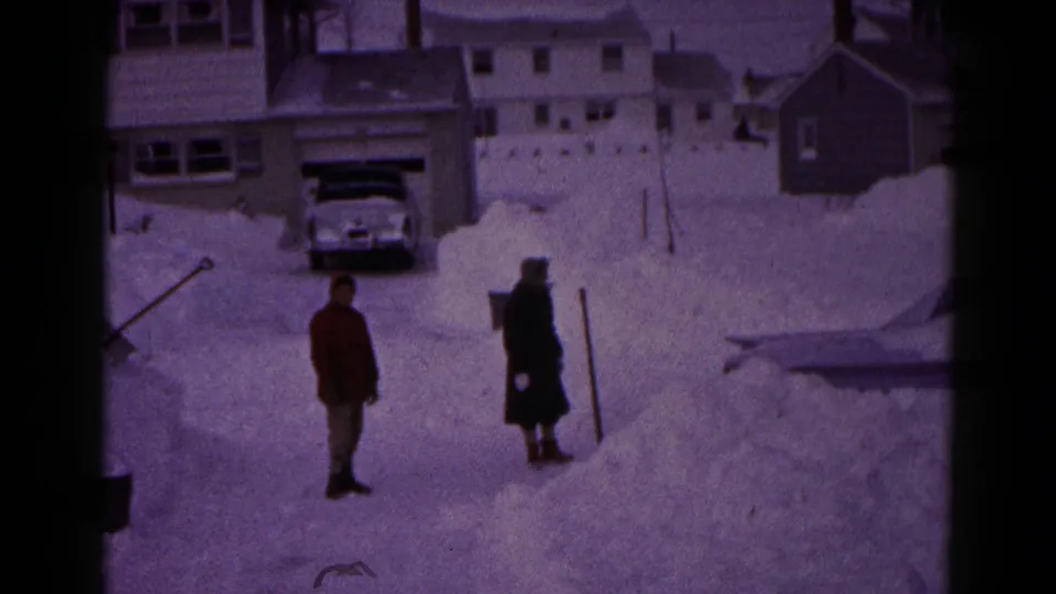 1959 Hagerstown frame showing two people in deep snow near a driveway, garage opening, shovel and parked vehicle area.