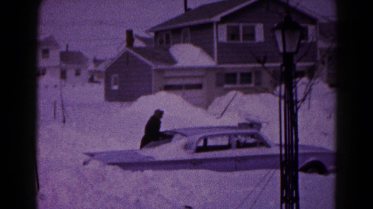 In 1959 Hagerstown, Maryland, a person stands on or beside a snow-covered sedan in a residential area with houses and a lamppost.