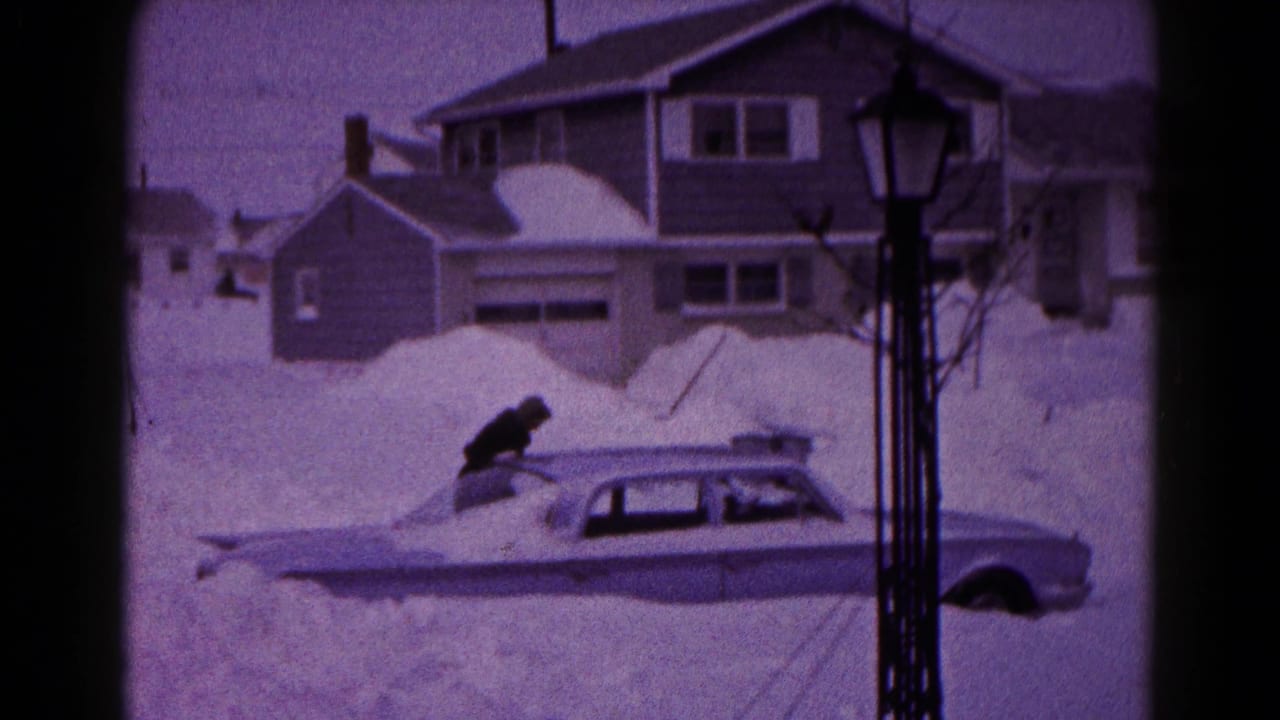 Person leans over the roof of a sedan buried in snow.