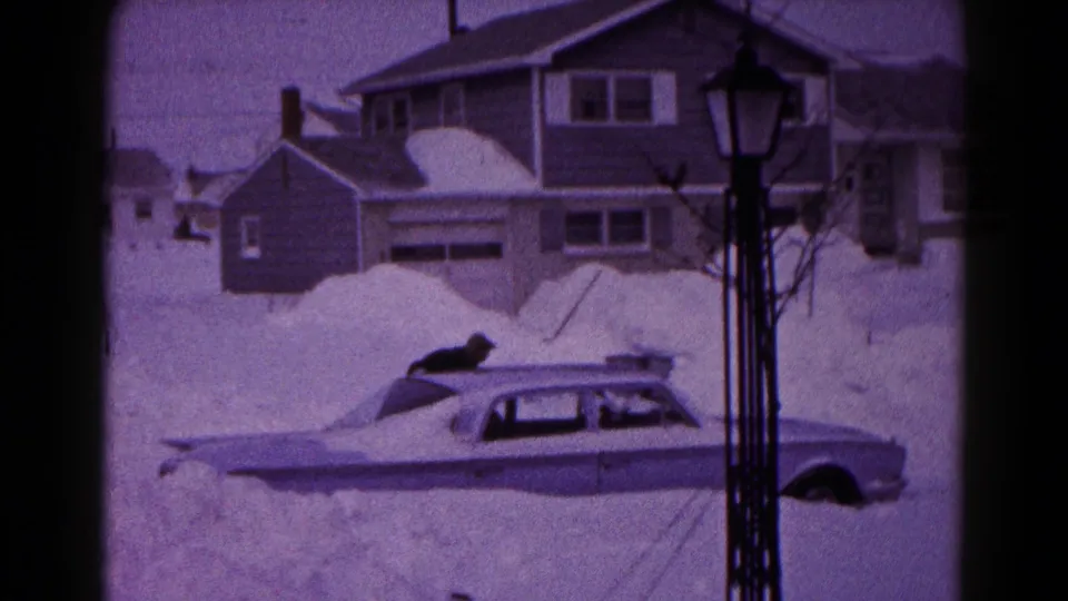 The person presses low against the roof of the buried sedan, with heavy snowbanks and a house visible in the background.