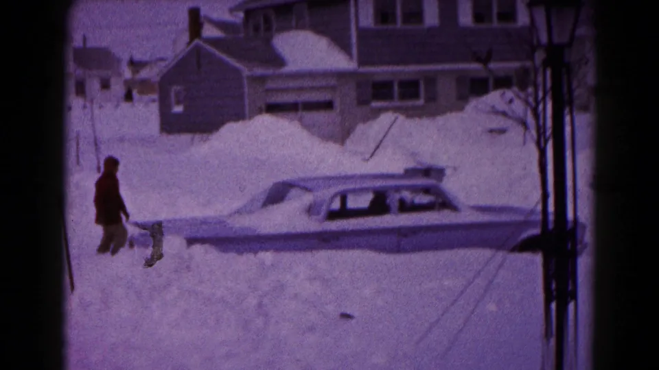 A person walks beside the partially buried sedan in the snowy Hagerstown residential scene, with houses and a lamppost nearby.