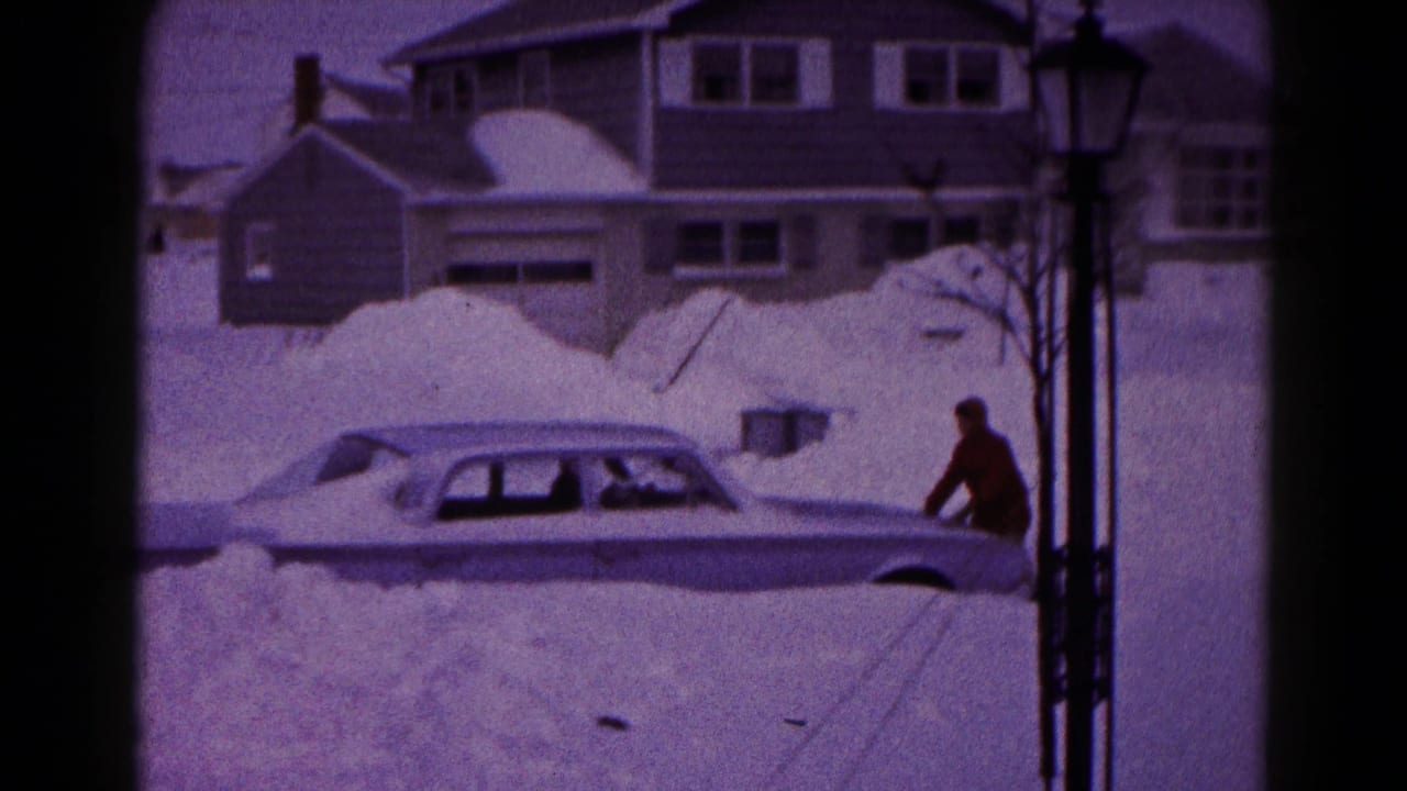Man stands near the front side of a sedan surrounded by heavy snow.