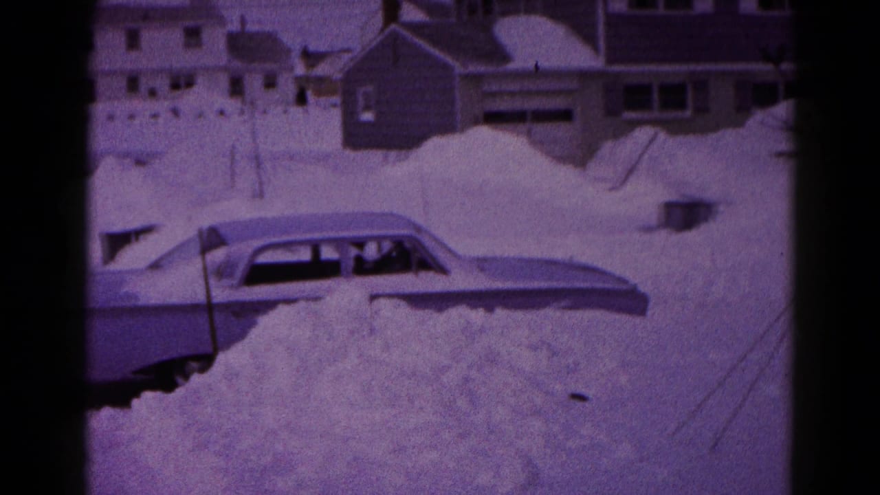 1959 Hagerstown home-movie frame showing a classic sedan nearly buried in deep snow beside residential houses.