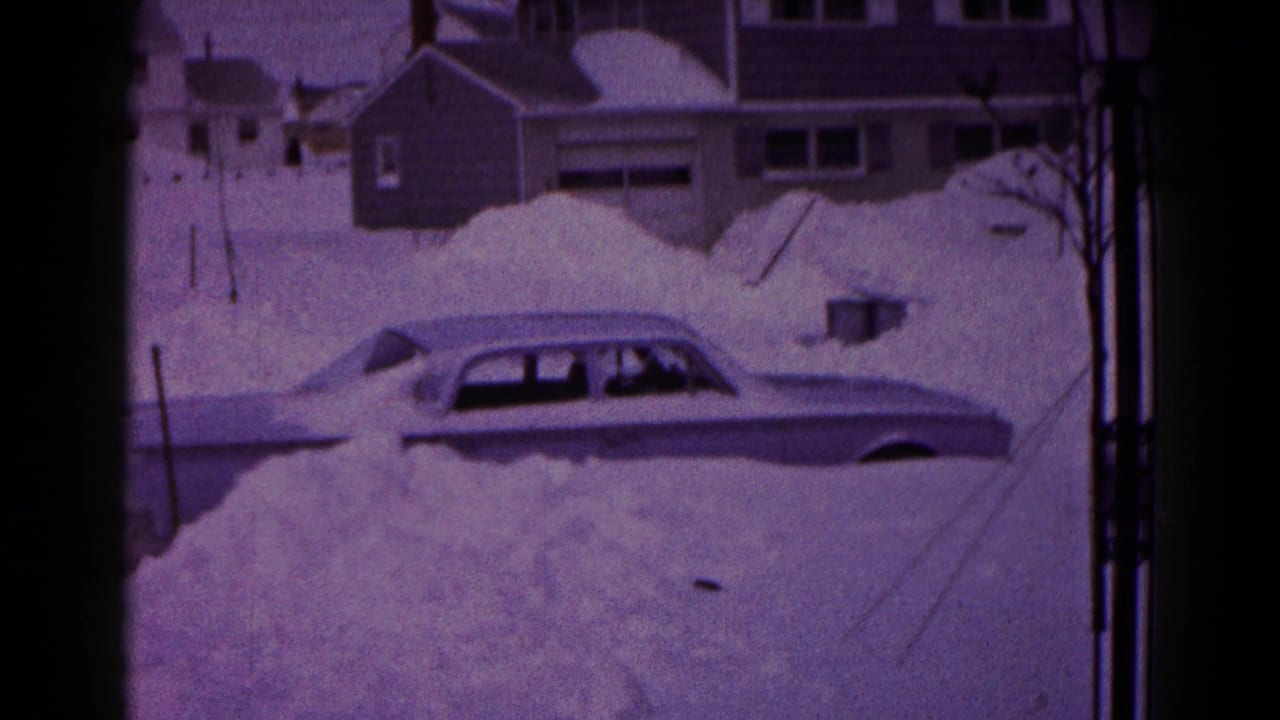 Snowbound sedan seen beside large drifts and neighborhood houses.